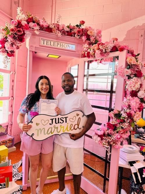 A couple stands in a pink phone booth, holding a "Just Married" sign and a marriage certificate, surrounded by flowers.