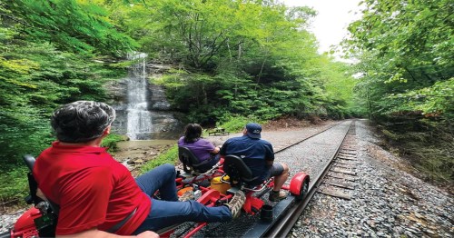Two people ride a rail bike along a track beside a lush forest and a waterfall in the background.