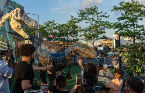 A crowd of people interacts with a life-sized dinosaur model at an outdoor event, with trees and attractions in the background.