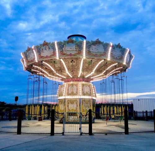 A brightly lit carousel ride at dusk, surrounded by a fence and under a cloudy sky.