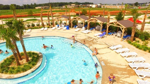 A sunny pool area with palm trees, cabanas, and people swimming and relaxing around the water.