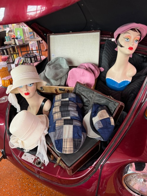 A vintage car trunk filled with hats and a suitcase, featuring mannequin heads displaying stylish hats.