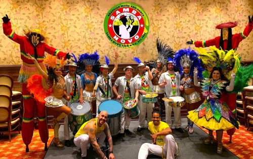 A vibrant group of performers in colorful costumes poses with drums, celebrating samba music and dance.