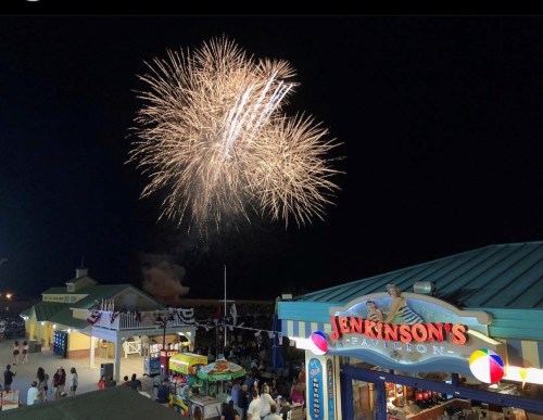 Colorful fireworks illuminate the night sky above a festive boardwalk scene with food stalls and a pavilion.