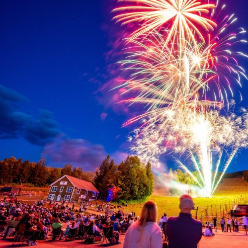 A crowd watches a vibrant fireworks display over a hillside, with a building in the foreground under a twilight sky.