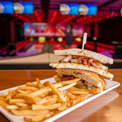 A stacked sandwich with chicken and peppers, served with a side of fries, in a vibrant bowling alley setting.