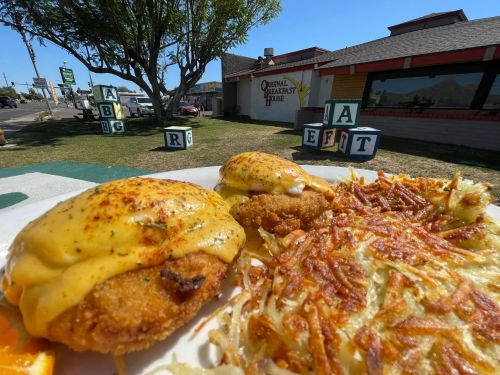 A plate of crispy fried food topped with cheese sauce, served with hash browns and an orange slice, outside a restaurant.
