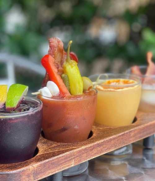 A wooden flight board with four colorful cocktails, garnished with various vegetables and fruits, set against a blurred background.