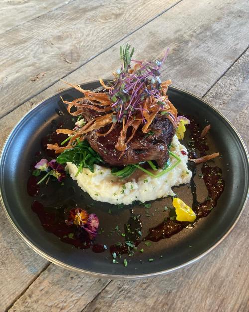 A gourmet dish featuring a meat cut on creamy mashed potatoes, garnished with crispy onions and edible flowers.