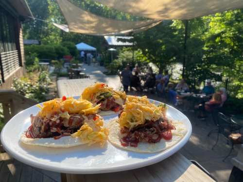 Three tacos topped with shredded cheese and sauce, served on a plate with a lively outdoor dining scene in the background.