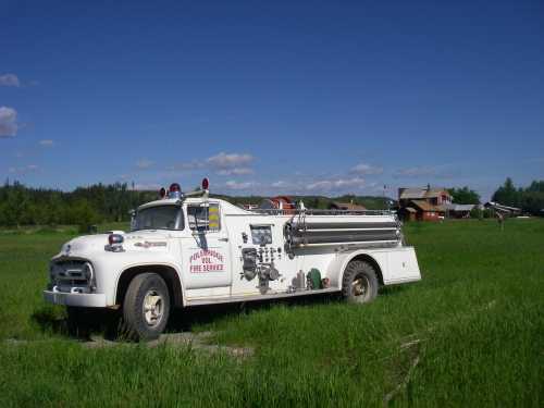 An old fire truck parked in a grassy field, with a blue sky and distant buildings in the background.