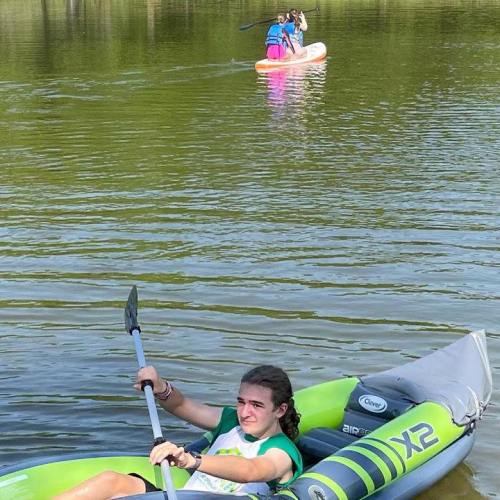 A girl paddles in a green kayak on a calm lake, while two others paddle on a pink inflatable boat in the background.