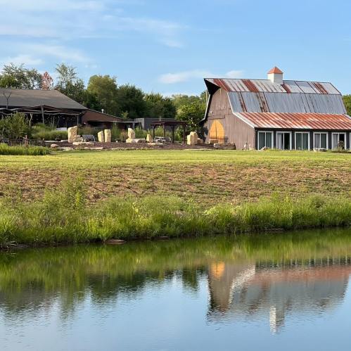 A rustic barn with a metal roof beside a pond, surrounded by green fields and trees under a clear blue sky.