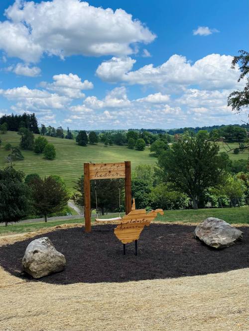 A wooden swing and sign in a grassy area, with trees and a blue sky in the background.