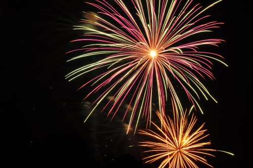 Colorful fireworks burst in the night sky, featuring pink, green, and orange trails against a dark background.