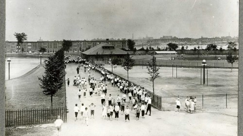 A historic black-and-white photo of a large group of people walking along a path in a park, with buildings in the background.