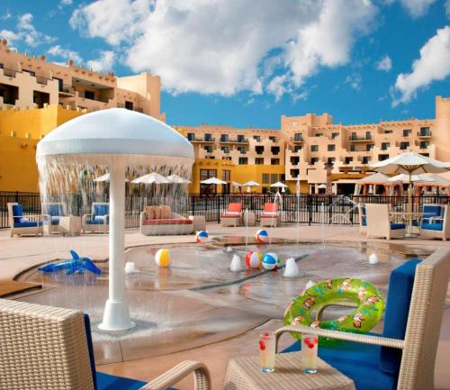 A vibrant pool area with water features, beach balls, and lounge chairs under a blue sky with fluffy clouds.