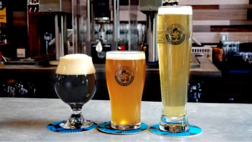 Three glasses of beer on a bar: a dark stout, a golden ale, and a tall pilsner, all with logo coasters.
