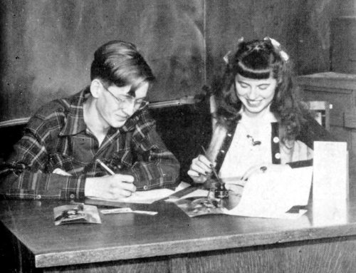 Two students, a boy and a girl, sit at a desk, smiling and working on assignments with papers and pens.