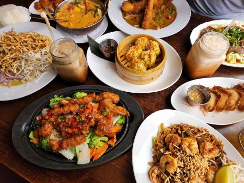 A spread of various Asian dishes, including stir-fried vegetables, shrimp, curry, and iced drinks on a wooden table.