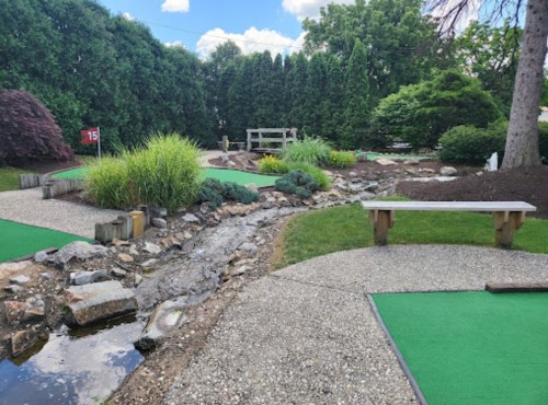 A mini-golf course featuring green turf, a rocky stream, and lush greenery under a blue sky with fluffy clouds.
