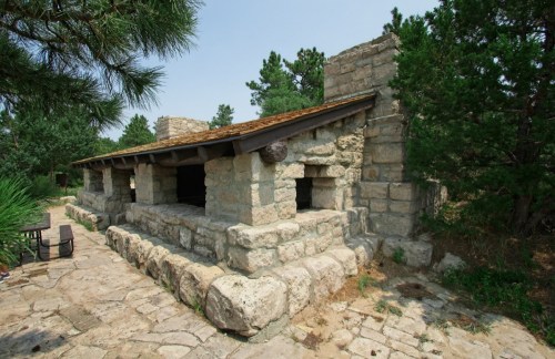 A rustic stone structure surrounded by trees, featuring a sloped roof and open spaces, set on a stone pathway.