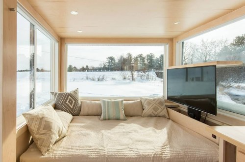 Cozy interior of a modern room with a large window, a bed with cushions, and a TV, overlooking a snowy landscape.