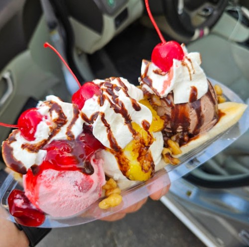 A hand holding a banana split with three scoops of ice cream, whipped cream, chocolate sauce, and cherries on top.