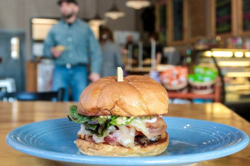 A close-up of a delicious burger with lettuce, bacon, and cheese on a blue plate, with a blurred background of a café.