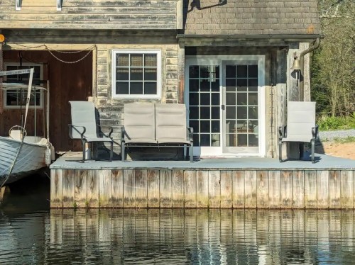 A wooden dock with two chairs facing a house, reflecting in calm water, with a boat nearby.