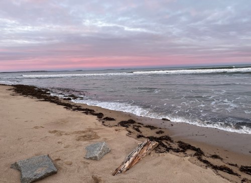 A serene beach scene at sunset, with gentle waves, sandy shore, and a pinkish sky.