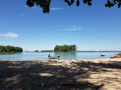 Two people near a calm lake on a sandy beach, with trees and a clear blue sky in the background.