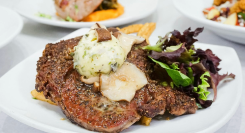 A plated steak topped with herb butter, mushrooms, and served with greens on the side.