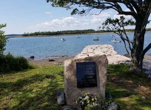 A stone monument by a calm waterway, with boats in the distance and trees framing the scene under a blue sky.