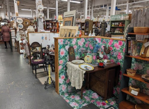 A vintage shop interior featuring floral wallpaper, antique furniture, and various collectibles on display.