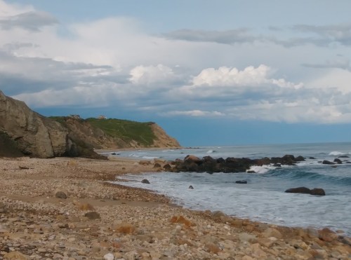 A rocky beach with gentle waves, cliffs in the background, and a cloudy sky above.