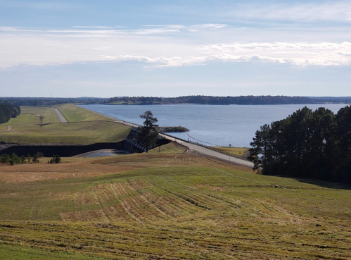 A scenic view of a dam with a road, surrounded by grassy fields and a calm lake under a clear blue sky.