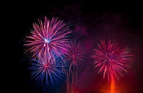 Colorful fireworks explode in the night sky, creating vibrant bursts of red, blue, and pink against a dark background.