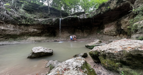 A group of people stands near a small waterfall in a rocky, forested area with a calm pool of water.
