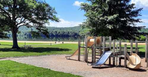 A playground with slides and climbing structures, set against a scenic lake and green hills under a blue sky.