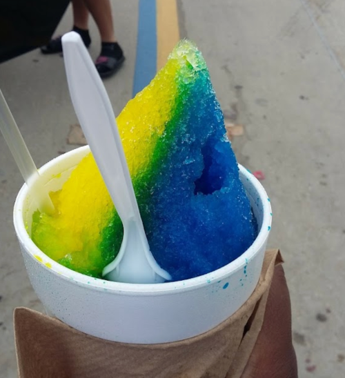 A cup of colorful shaved ice with blue, green, and yellow layers, held by a hand with a spoon.