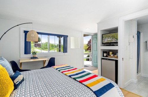 Cozy bedroom with a striped blanket, desk, and kitchenette, featuring bright natural light and a welcoming entrance.