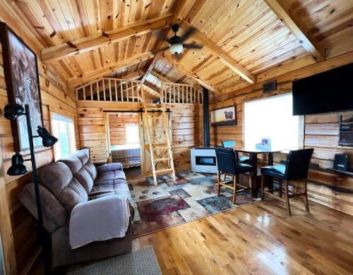 Cozy wooden cabin interior with a loft, sofa, dining area, and large windows, featuring warm wood tones and a rug.