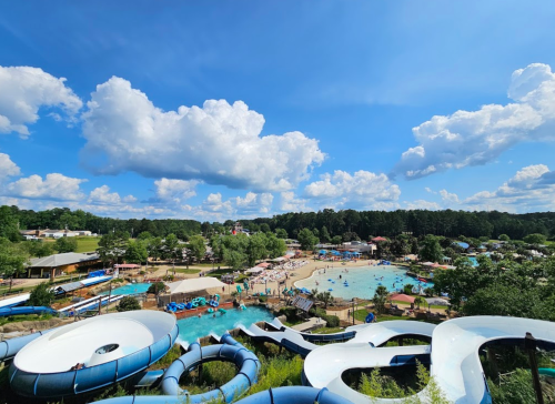 A vibrant water park scene with slides, pools, and people enjoying a sunny day under a blue sky with fluffy clouds.