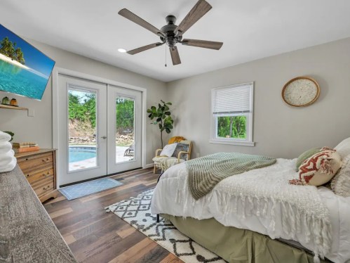 Cozy bedroom with a queen bed, ceiling fan, TV, and sliding doors leading to a pool area. Natural light and greenery.