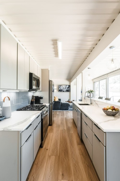 Modern kitchen with sleek cabinets, stainless steel appliances, and a view of a living area through large windows.