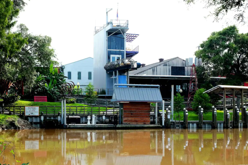 A riverside view featuring a modern building, dock, and lush greenery reflecting in the water.