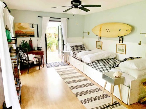 Cozy bedroom with two beds, surfboard on the wall, and a view of a garden through open doors. Light and airy decor.