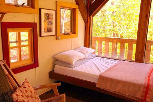 Cozy bedroom with a bed, wooden accents, and large windows overlooking a forested area. Warm, inviting atmosphere.