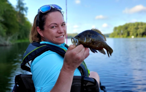 A woman in a blue shirt smiles while holding a fish, sitting by a calm lake on a sunny day.
